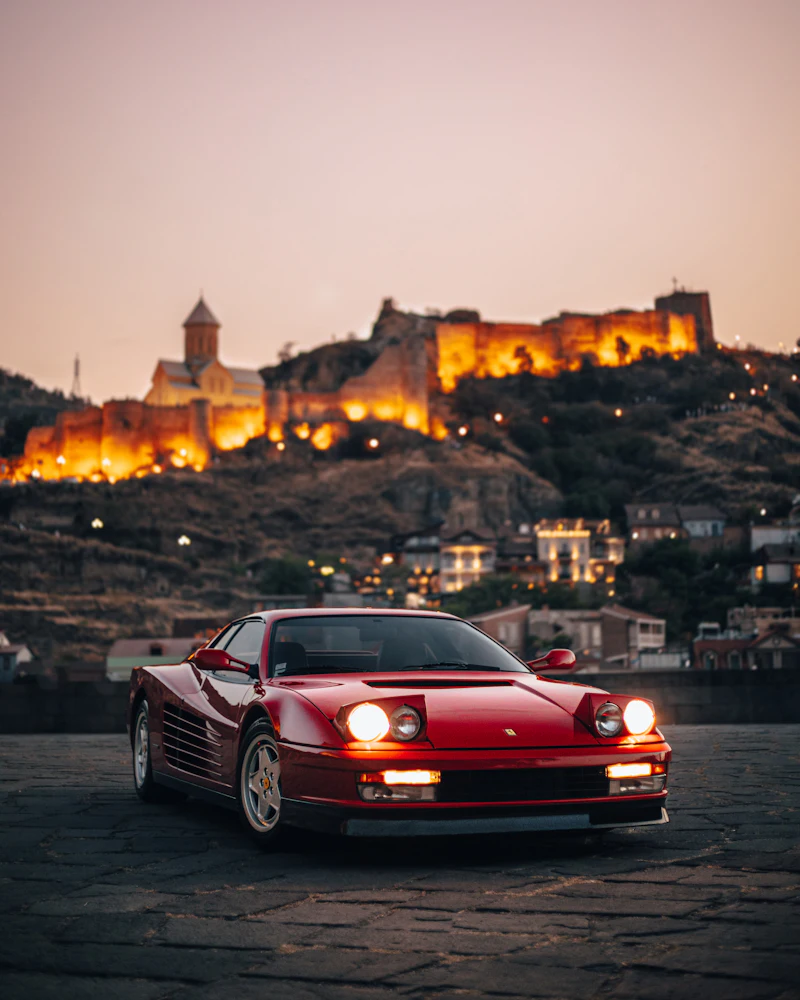 A red Ferrari Testarossa at dusk, with a lit hilltop fortress behind it