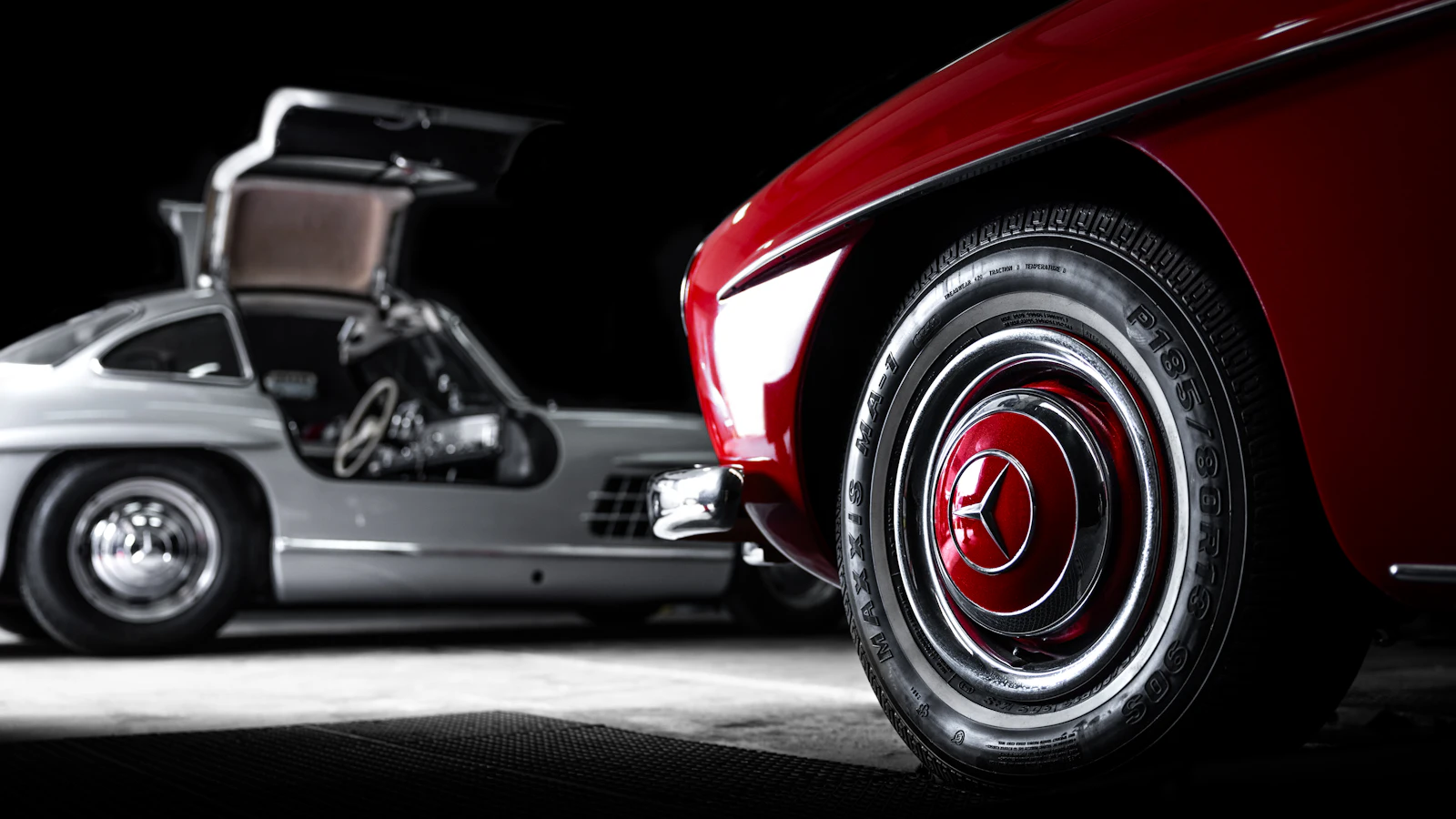 A silver Mercedes-Benz 300 SL Gullwing with its doors open, beside a red roadster in a dark studio